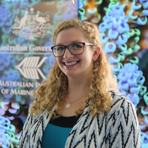 Woman with blonde curly hair wearing glasses, standing in front of a colourful background.