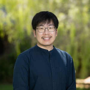 Smiling man with short black hair and glasses in navy blue long-sleeved shirt, standing in front of green tree. 