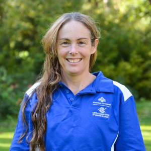 Smiling woman auburn hair wearing a royal blue and white shirt standing outside and is in front of green trees.