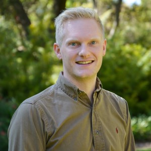 Man wearing long-sleeved olive shirt standing in front of a bushy green garden