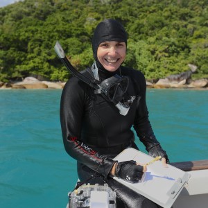 Woman in black diving suit, sitting on the edge of a boat holding clipboard and, in front of an ocean and green bushy mountain.