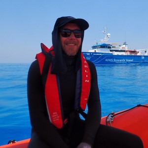 Man wearing black dive suit and black sunglasses, sitting on the edge of orange boat in the ocean with AIMS Research Vessel in the background.