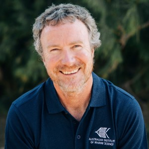 Photo of bearded man wearing navy blue polo, sitting in front of dark green garden.