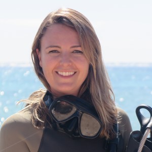 Woman, long hair, wearing scuba suit in front of ocean.