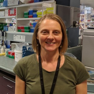 Woman, Short hair wearing green shirt. Standing in front of shelf. 