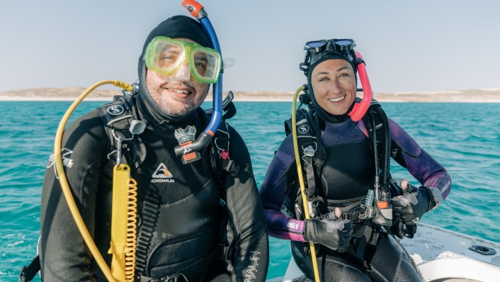 2 people sitting in dive gear on the edge of a small vessel smiling at camera