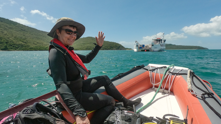 A woman sits on a small boat in a wetsuit and surrounded by scuba diving equipment