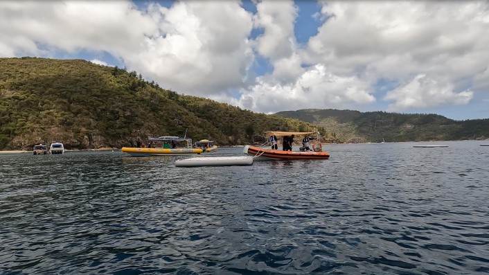 a wide photo of a calm sea with a green hill in the background. There are a few small boats on the water