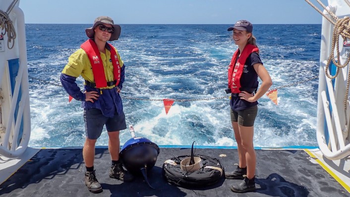 Two people standing on the deck of a boat. At their feet is an array of ropes and equipment