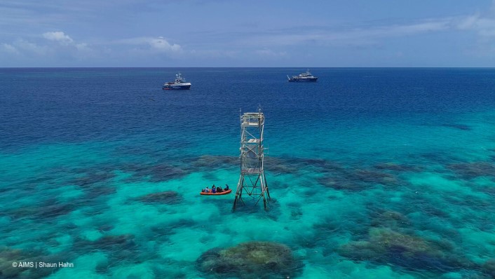 An aerial viewo of a coral reef with a metal structure on the reef and two vessels in the distance