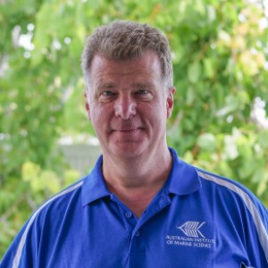 Man wearing blue shirt, standing in front of green trees.