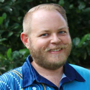 Bearded Man wearing blue shirt standing in front of green leaves.