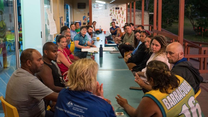 many people sitting around a long rectangular table on an outside deck