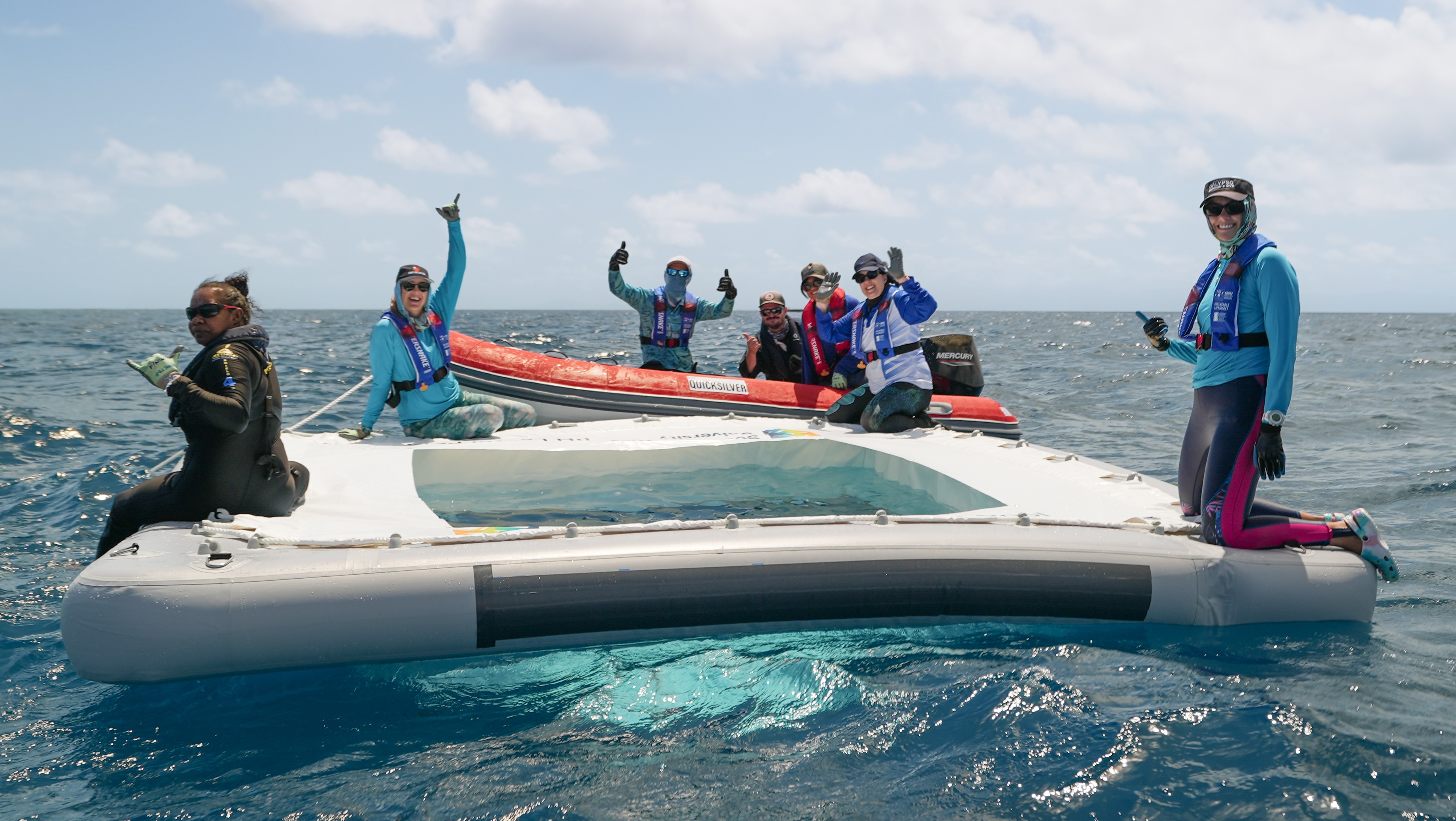 people sitting in a small boat and on the edge of an inflateable square, waving at the camera.