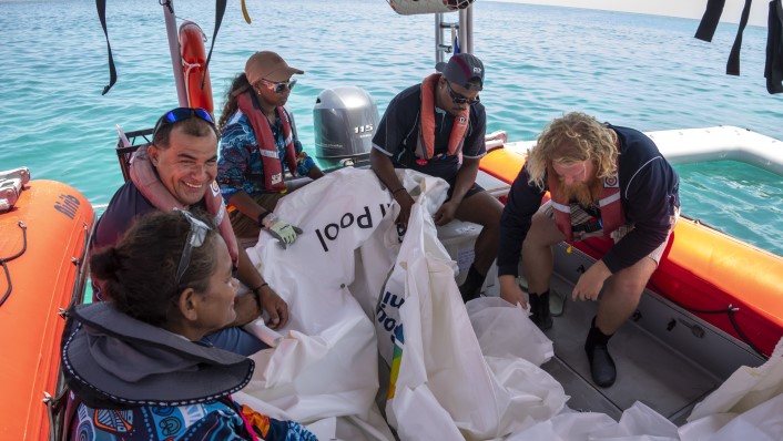 many people in a small boat on the water with large vinyl sheets, clearly deflated.