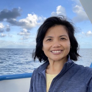 Woman with brown hair wearing blue jacket with ocean in the background