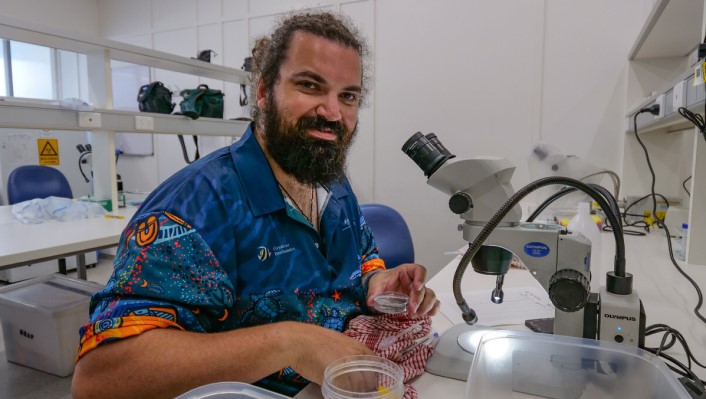 A man in front of a microscope looks at the camera and smiles