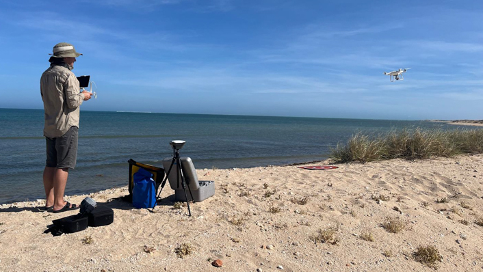 person standing on a beach with bags at their feet, looking at a small drone about 3m above the sand