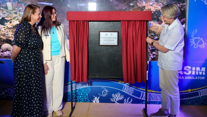 three people standing in front of a large colourful aquarium having just revealed a plaque which is between some red curtains