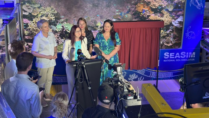 people standing at lecturn in front of large colourful aquarium with red curtains to the side. They face a number of people with cameras and lights.