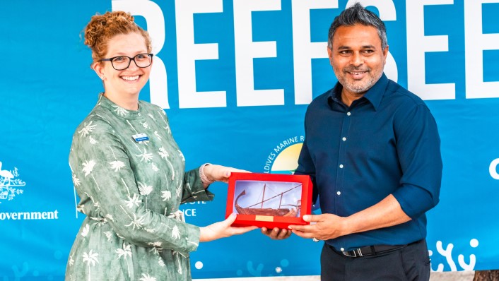 two people smiling while both holding a gift exchange. A ReefSeed banner is behind them