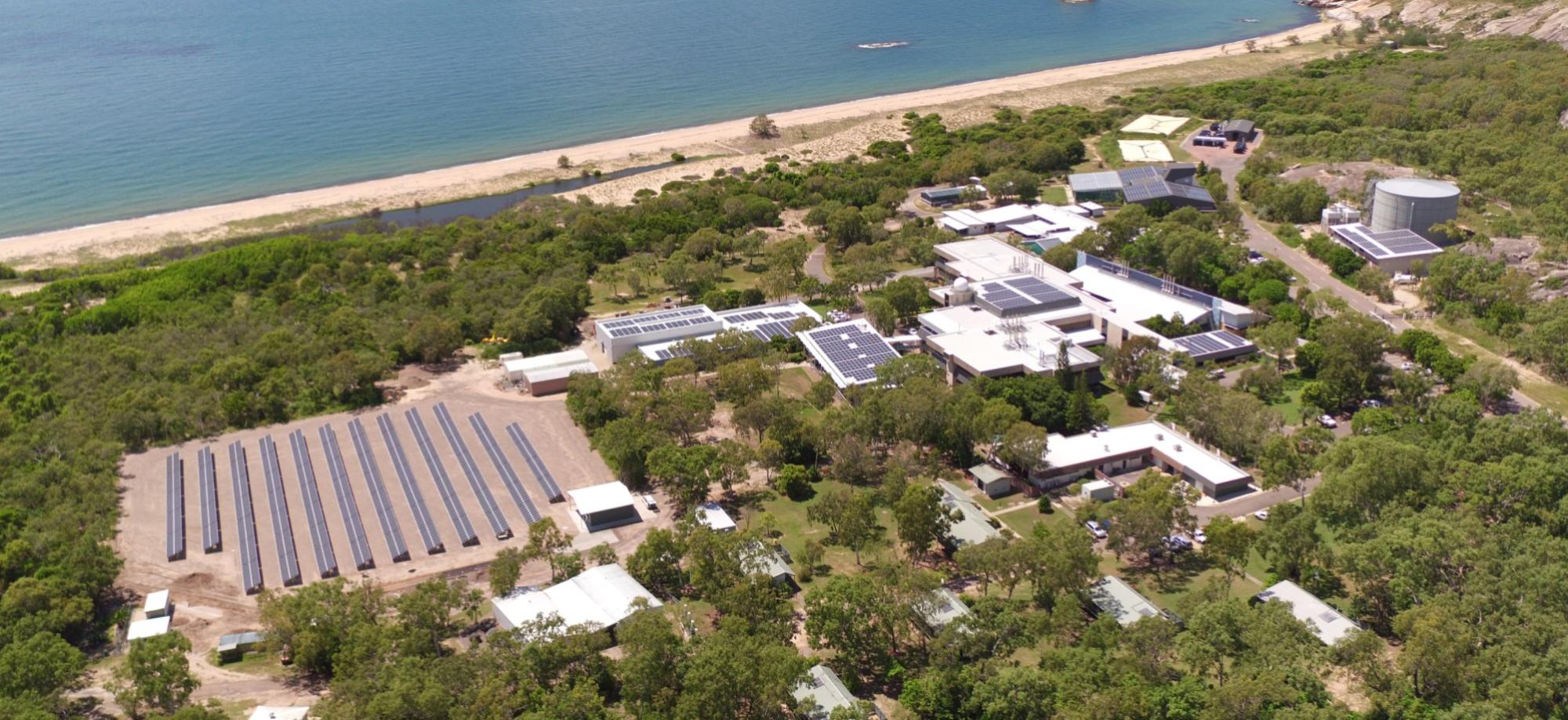 This photo is taken from the air. Buildings with solar panels are surround be green grass and bush. The ocean and beach can be seen at the top of the photo. 