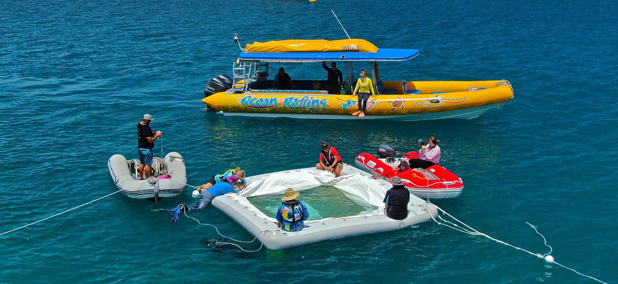 Awhite floating square lies on the surface of the ocean with a yellow boat behind it and grey bot beside it. There are people on the boats and on the floating square. The water is calm and it's a sunny day. 