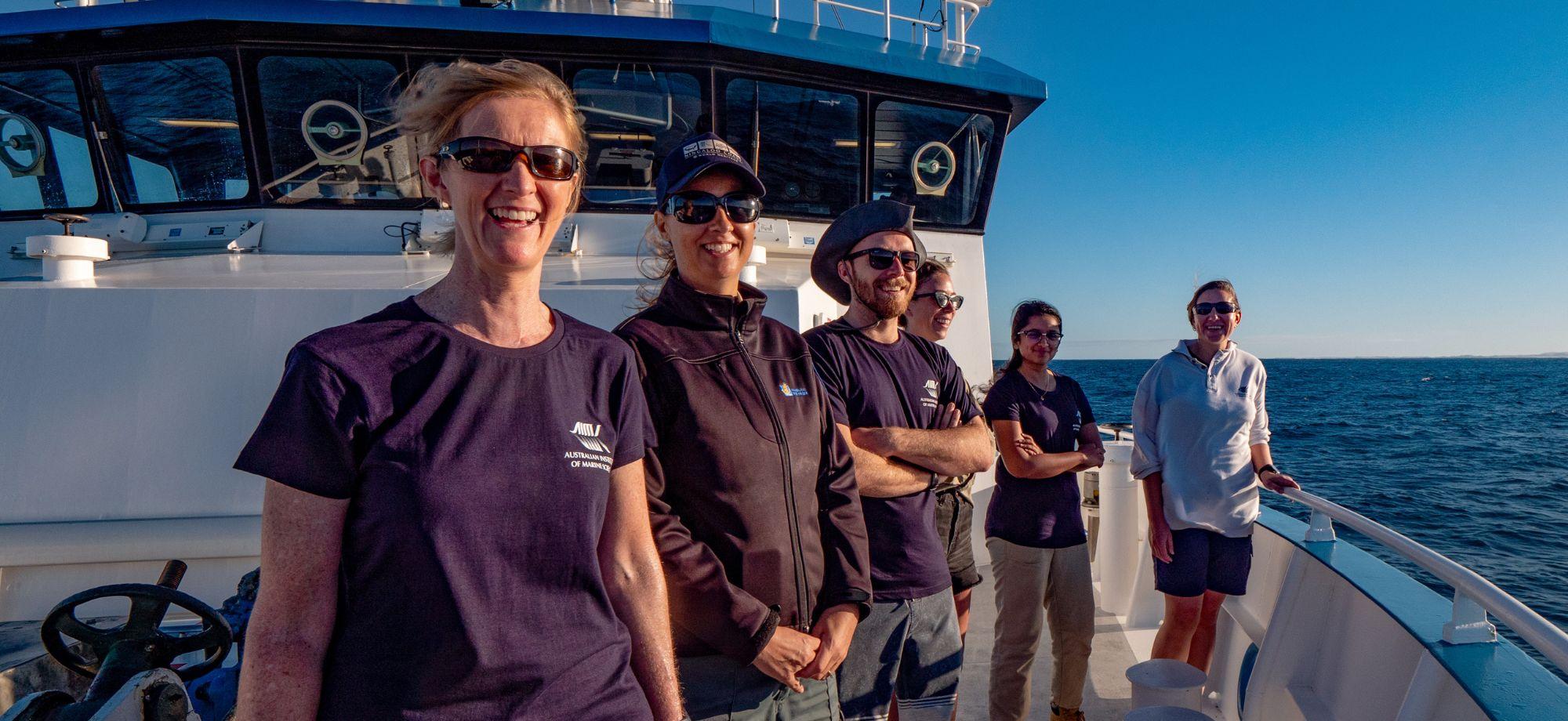 five people in AIMS shorts smiling while standing on the bow of the RV Solander with a calm sea in the background