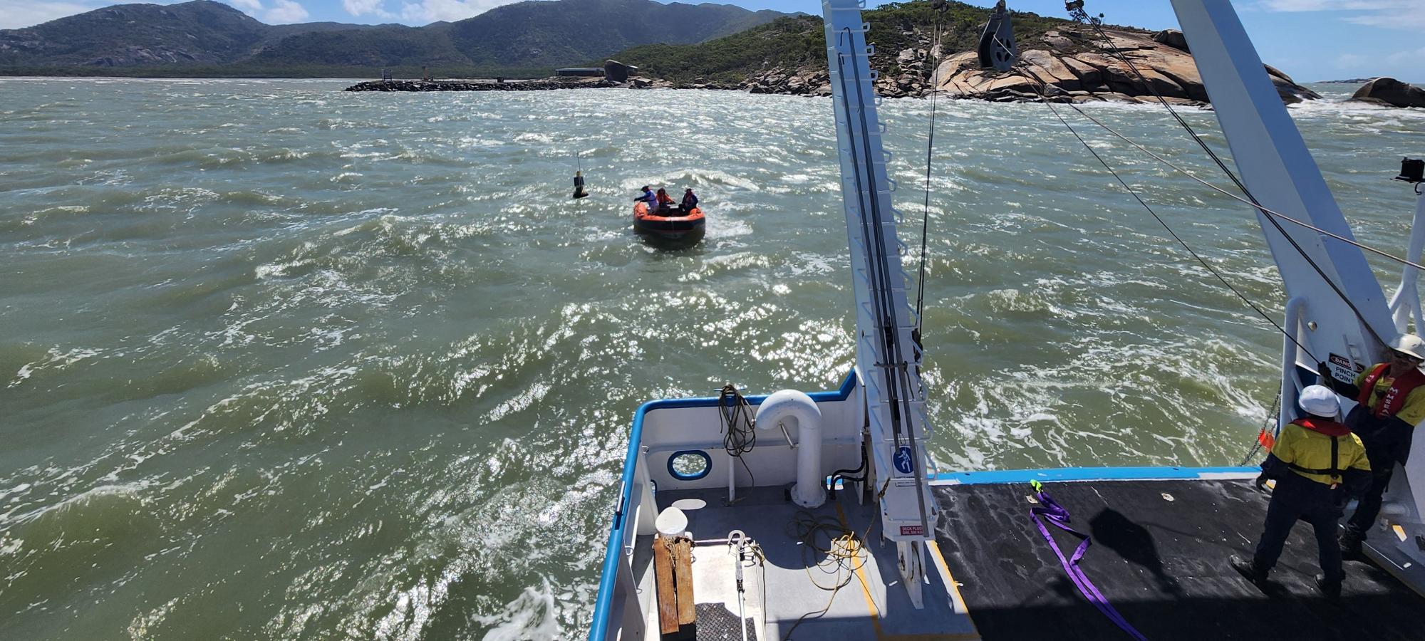 a view over the stern of a vessel at small orange inflatable in the water, and a green, hilly coastline in the distance