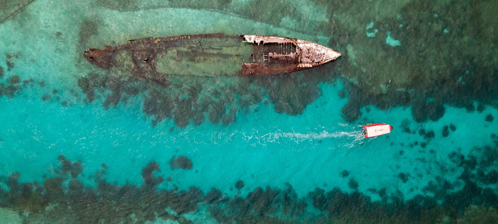 Overhead view of a small boat motoring through a turquoise channel next to a ship wreck