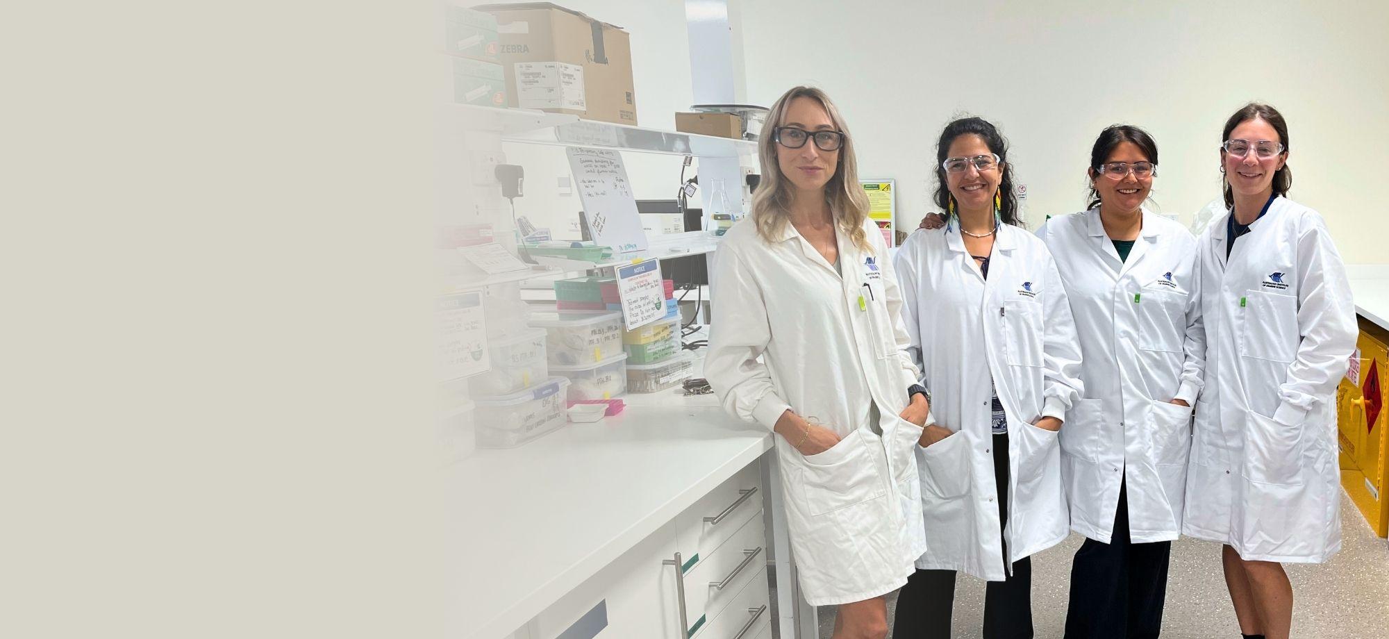 4 women in lab coats and safety glass standing in a line, smiling at the camera in a lab with equipment