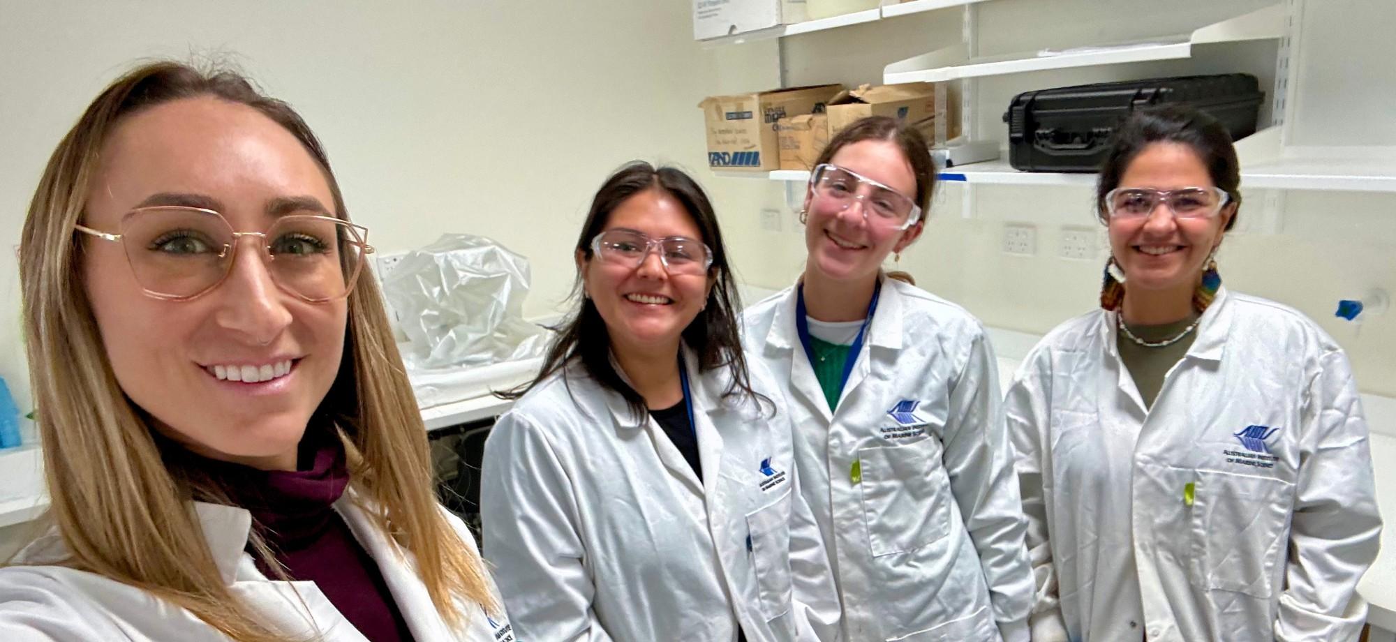 4 women in lab coats and safety glasses smiling at the camera in a selfie