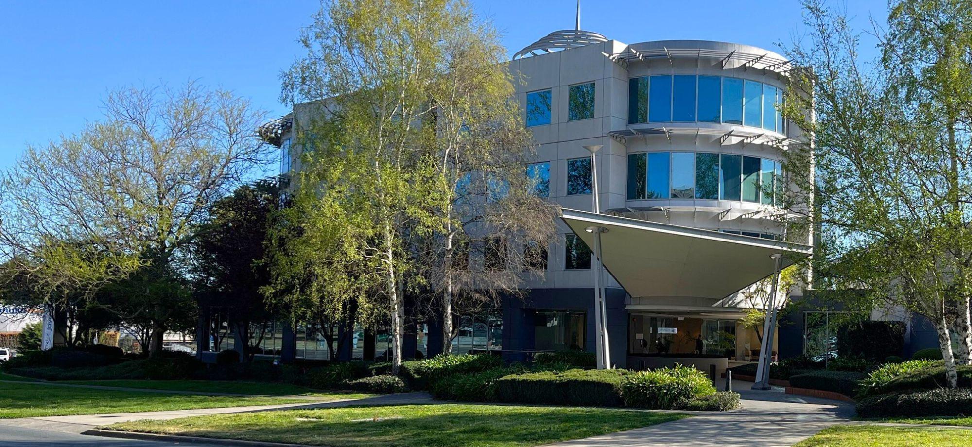 photo of modern building surrounded by green grass and tall trees. the sky is clear.