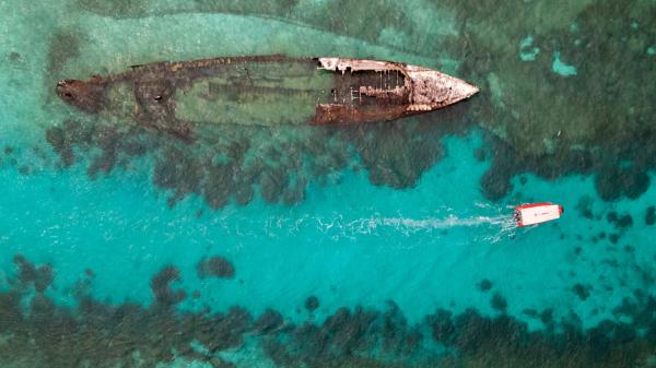 Overhead view of a small boat motoring through a turquoise channel next to a ship wreck