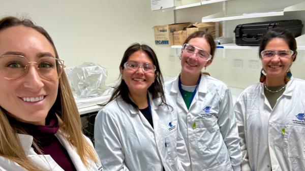 4 women in lab coats and safety glasses smiling at the camera in a selfie