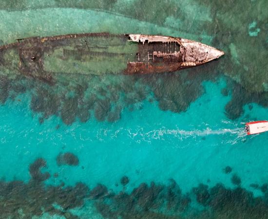 Overhead view of a small boat motoring through a turquoise channel next to a ship wreck