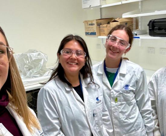 4 women in lab coats and safety glasses smiling at the camera in a selfie