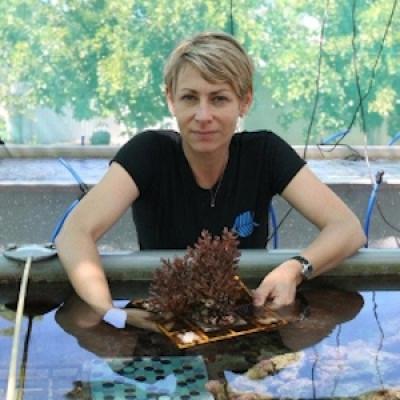 Woman with short blonde hair holding coral over water, with green tree in the background