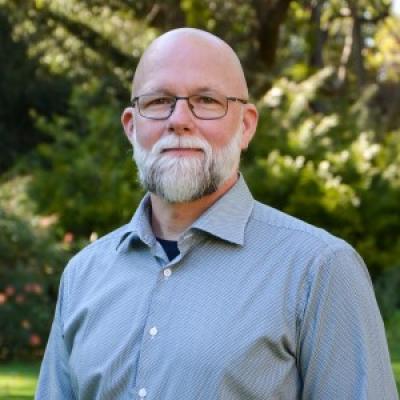 White bearded man with glasses wearing long-sleeved light blue dress shirt, standing outside in front of green trees.